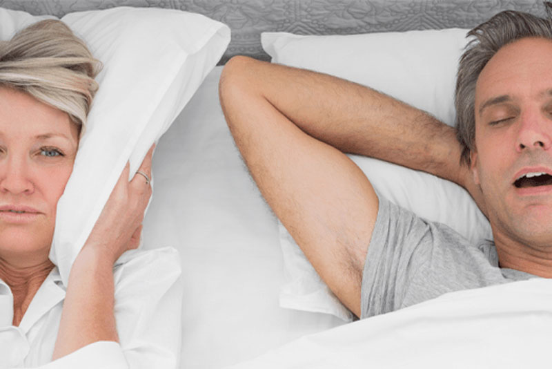 A woman covers her ears with a pillow in bed as she lays next to a man who is snoring loudly.